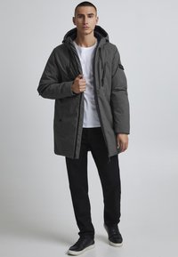 Parka gris matelassée avec capuche, fermeture éclair à l'avant, deux poches latérales et un accent noir sur la manche. Portée par-dessus un t-shirt blanc et un pantalon noir.