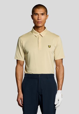 Homme portant un polo beige avec un logo d'oiseau jaune, un pantalon bleu marine, et un gant de golf blanc à la main droite, sur fond gris.