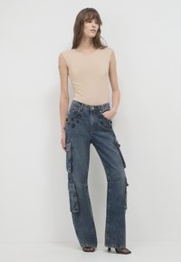 Body color beige aderente abbinato a pantaloni cargo in denim scuri a vita alta e larghi, dotati di molteplici tasche e dettagli decorativi neri.