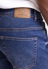 Mørkeblå denimjeans med teksturert overflate. Med en synlig brun lærmerkelapp preget med "REDEFINED REBEL" og oransje søm.