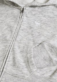 Sweat à fermeture éclair gris en tissu doux chiné, avec une poche avant et un petit logo brodé blanc sur le côté gauche.