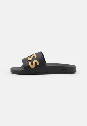 Sort klipklap med bred rem med store hævede gyldne bogstaver og tekstureret sål, vist på hvid baggrund.