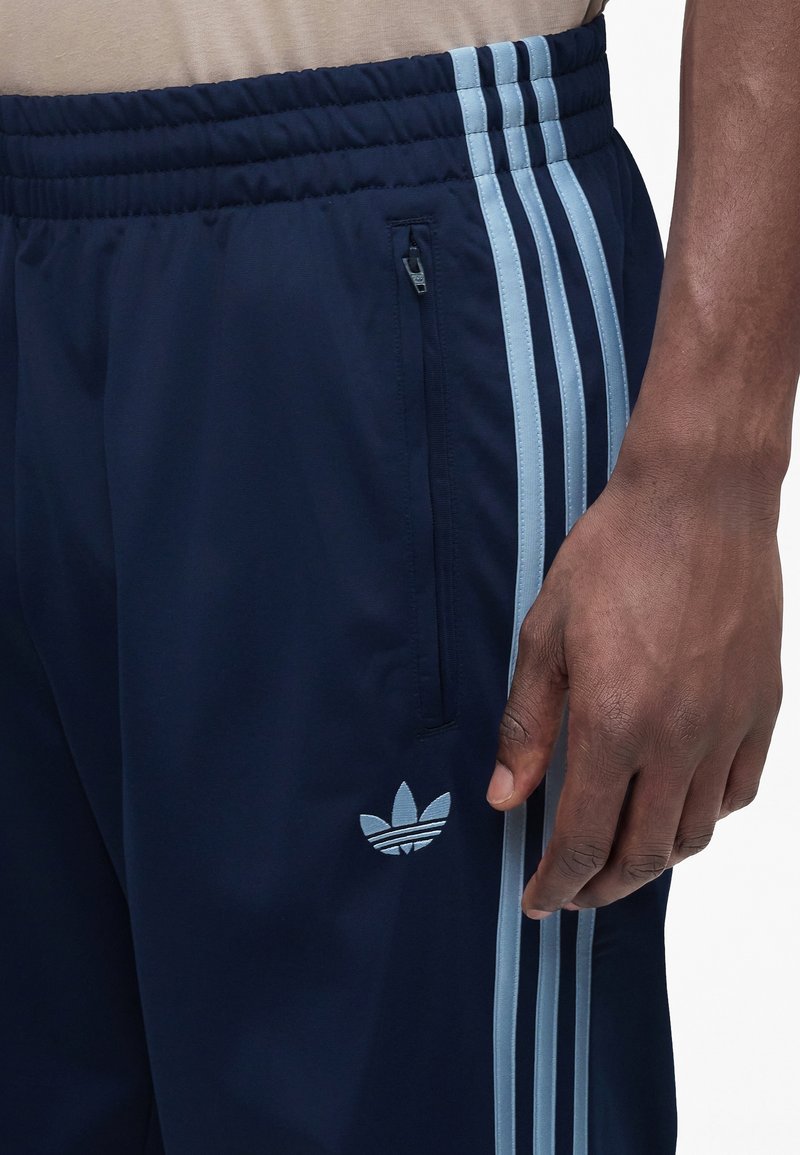 Close-up of a person wearing navy blue Adidas track pants with light blue stripes, zipper pocket, and Adidas logo near the hip.