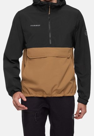 Homme portant un anorak à capuche Mammut noir et marron avec une fermeture éclair frontale et une grande poche à rabat, associé à un pantalon noir.