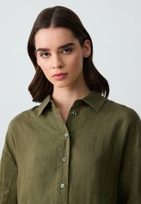 Camicia in lino verde oliva con colletto, sette bottoni e vestibilità comoda, che mostra una sottile texture e pieghe naturali.