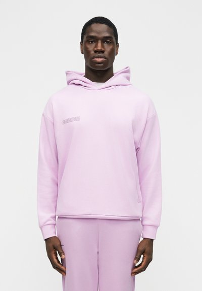 Fiatal férfi világoslila kapucnis pulóverben és hozzáillő nadrágban, karjait lazán tartva, egy egyszerű fehér háttér előtt áll.