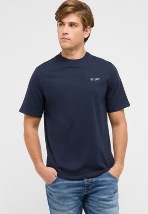 Młody mężczyzna ubrany w granatową koszulkę Mustang i niebieskie dżinsy, stojący z rękami w kieszeniach, patrzący lekko na bok.