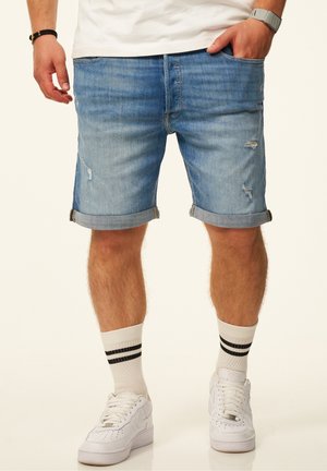 Mann trägt hellblaue, hochgekrempelte Jeansshorts, weiße Socken mit schwarzen Streifen, weiße Sneakers, ein weißes T-Shirt und Armband-Accessoires.