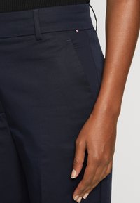 Pantaloni tailored de culoare bleumarin, confecționați dintr-un material neted, cu buzunare laterale și un mic logo cu accente roșii și albe aproape de talie.