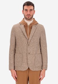Uomo che indossa un blazer a quadretti beige sopra un hoodie tan con zip e pantaloni color cammello, in piedi con le braccia rilassate ai lati.