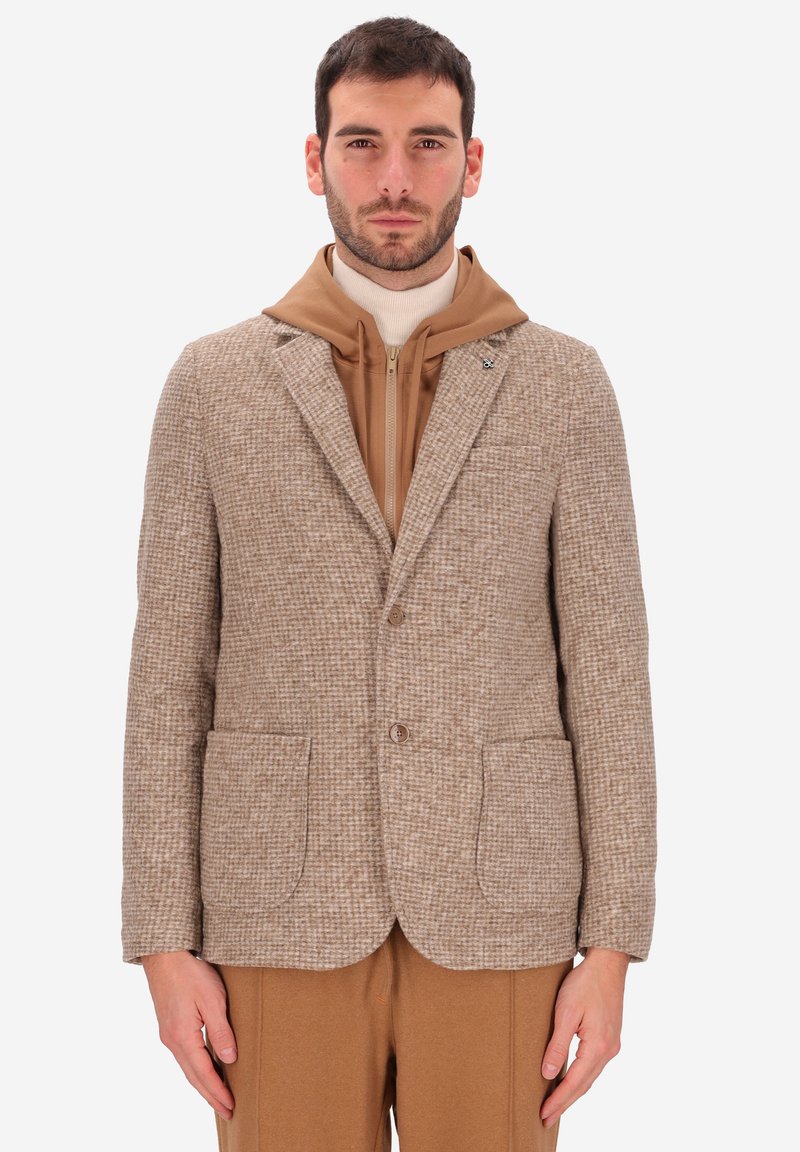 Uomo che indossa un blazer a quadretti beige sopra un hoodie tan con zip e pantaloni color cammello, in piedi con le braccia rilassate ai lati.