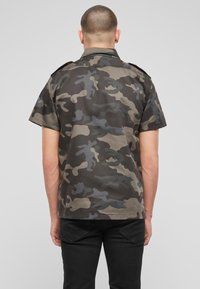 Camicia camouflage in tonalità verde scuro e grigio, con maniche corte, chiusura a bottoni e spalline sulle spalle, texture liscia e orlo dritto.