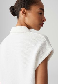 Indumento bianco strutturato con maniche corte e colletto, caratterizzato da un morbido motivo intrecciato visibile sulla superficie.