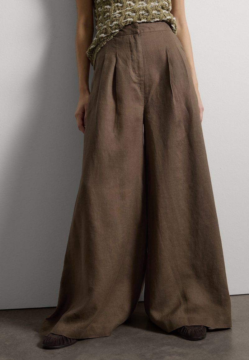 Hochtaillierte, weit geschnittene braune Leinenhose, kombiniert mit dunklen flachen Schuhen, getragen zu einem strukturierten, ärmellosen Stricktop.