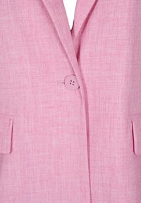 Gros plan sur le devant d'un blazer rose montrant un tissu texturé, un seul bouton fermé et deux poches à rabat de chaque côté.