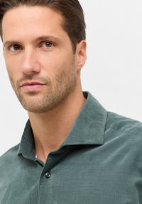 Camicia in corduroy grigio-verde con colletto a bottoni, caratterizzata da un tessuto texturizzato e un singolo bottone nero nella parte anteriore.