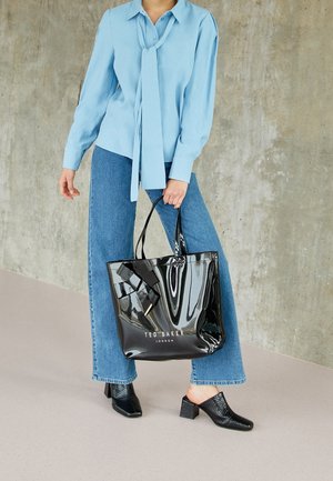 Musta kiiltävä käsilaukku, jossa on rusettikoriste ja etupuolella kohopainettu logo, yhdistetty vaaleansiniseen puseroon ja leveälahkeisiin farkkuihin. Teksturoidut mustat kengät.