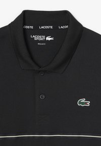 Camiseta tipo polo negra con acabado texturizado. Cuenta con un cuello negro, dos botones, marca en blanco y un logo de cocodrilo verde en el pecho.