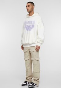 Overdimensioneret cremefarvet hoodie med et lilla sommerfugle-grafik og lommer foran, parret med beige cargo bukser med flere lommer og sorte sneakers.