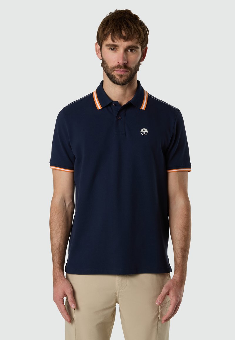 Hombre con camisa polo azul marino con cuello y bordes de las mangas con rayas naranjas, de pie contra un fondo claro y liso.