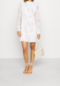 White floral dress with long sleeves, textured lace accents, and a flared hem. Paired with strappy white sandals and a woven purse.