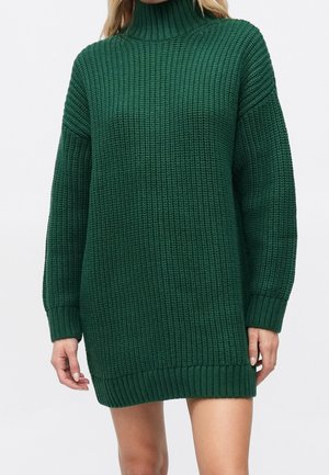 Kobieta w długiej, ciemnozielonej, grubym dzianym sukience sweterkowej z golfem i długimi rękawami, stojąca na jednolitym tle.