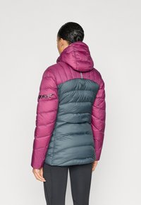 Μπουφάν με επένδυση, με μοβ κουκούλα και ώμους, και πετρόλ σώμα. Διαθέτει λογότυπο στην πλάτη και υφασμάτινη υφή με σχεδιασμό καπιτονέ.