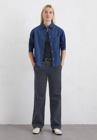 Blaue Denim-Hemd mit langen Ärmeln, knopfbarer Vorderseite, einzelner Brusttasche; kombiniert mit lockeren grauen Hosen und hellen Schuhen.