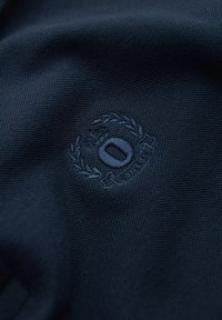 Marinfärgad tyg med en strukturerad yta som har en broderad logotyp omgiven av lagerblad och siffran noll.