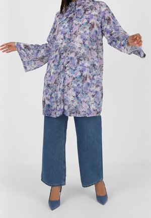 Woman wearing a long-sleeve, floral purple blouse with wide cuffs, paired with wide-leg blue jeans and blue pointed heels.