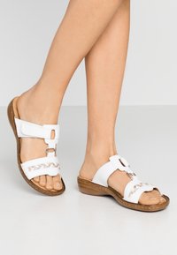 Sandales blanches à semelle plate avec un dessus texturé, présentant un design tressé et des accents métalliques. La semelle est brun clair avec un léger talon.