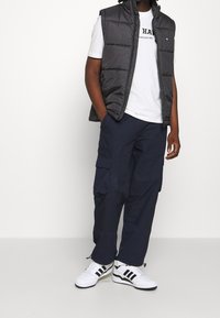 Černá vesta s polstrováním přes bílé tričko s logem, doplněná tmavě modrými kalhotami typu cargo a bílými botami s černými pruhy.