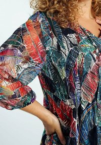 Kihara henkilö, joka pukeutuu läpikuultavaan puseroon, jossa on värikäs, abstrakti höyhenkuvio ja piispa-hihat, käsi lantiolla.