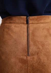 Falda de ante marrón con una superficie texturizada, que cuenta con una cremallera en la parte trasera con un tirador metálico y un diseño ajustado que enfatiza la cintura.