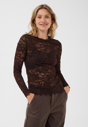 Femme souriante, portant un haut en dentelle marron foncé à manches longues et un pantalon marron avec les mains dans les poches, debout devant un fond uni clair.