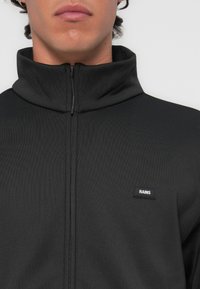 Black zip-up jacket with a high collar, ribbed texture, and a small logo label on the left side. Features a sleek, minimalist design.