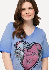 Top azul de manga corta con cuello en V que presenta un diseño de corazón, ilustraciones de ciervos, acentos florales y texto en un tono rosa contrastante.