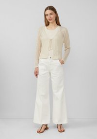 Helles beiges Strickcardigan mit offenem Vorderteil und Bindedetail, kombiniert mit weißen weit geschnittenen Hosen und braunen Sandalen mit einem gemusterten Riemen.