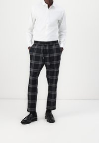 Weißes Hemd mit Knopfleiste, kombiniert mit grau-schwarz karierten Hosen mit elastischem Bund und Taschen. Schwarze Schuhe ergänzen das Outfit.