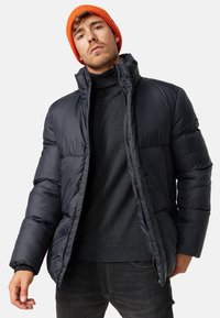 Mørkeblå pufferjakke med høj krave og lynlås, parret med en mørkegrå sweater og en orange strikket hue. Let og isoleret.