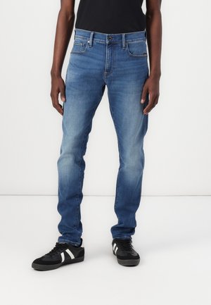 Mann trägt ausgewaschene blaue Slim-Fit-Jeans und schwarze Sneaker mit weißen Streifen, steht vor einem weißen Hintergrund.
