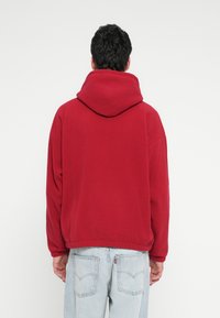 Felpa rossa oversize in tessuto morbido con cappuccio ampio. Presenta maniche larghe e orlo corto, abbinata a jeans azzurri.