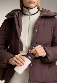 Isoleret pink vandflaske med brun låg holdt i hånden, iført en mørk burgunder puffjakke og en lysfarvet zip-up top.