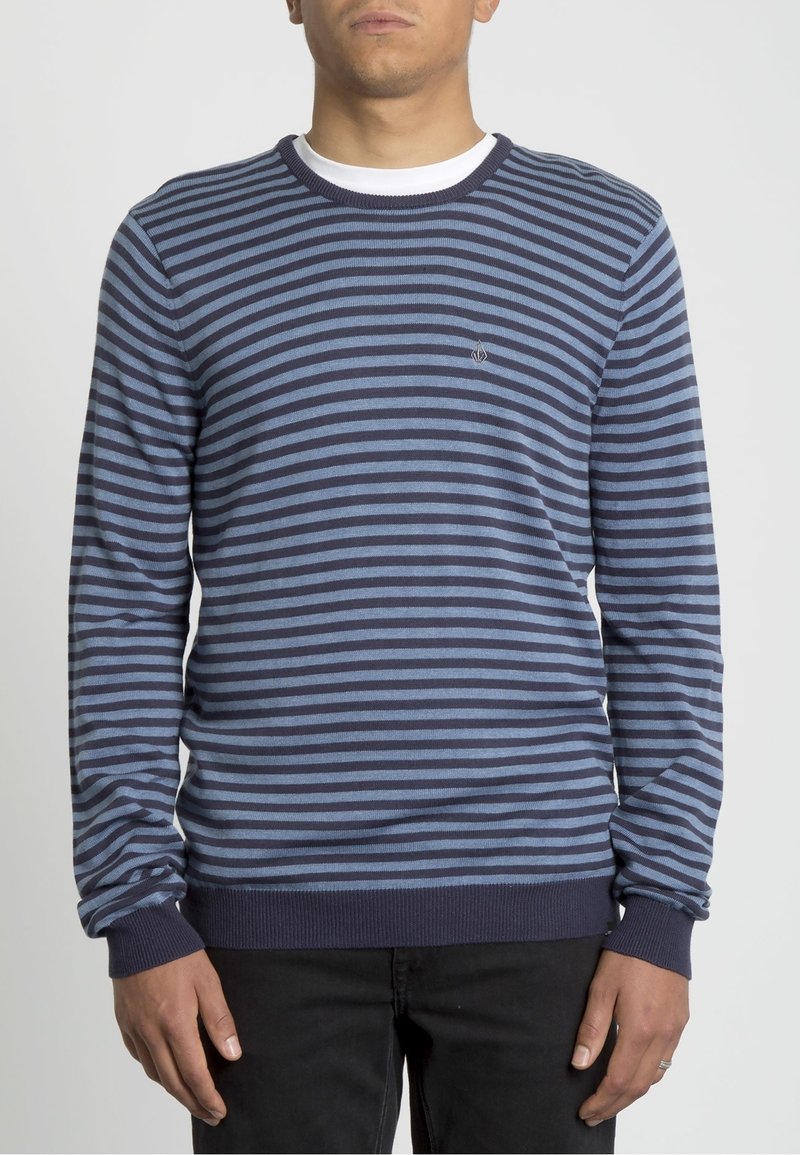 Homme portant un pull à rayures horizontales bleu marine et bleu clair sur une chemise blanche, associé à un pantalon noir, debout devant un fond uni.
