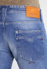 Jeans de mezclilla azul con un ligero efecto desgastado, que cuentan con un parche de cuero marrón etiquetado "REPLAY" y bolsillos traseros detallados con acentos de costura.