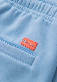 Pantalons de survêtement bleu clair avec taille élastique, présentant une patche rectangulaire orange portant l'inscription "LEVEL UP". Texture douce, design minimaliste.
