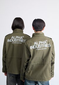 Olíva zöld kabátok fehér "ALPHA INDUSTRIES STUDIO EDITION" felirattal a háton. Gallérral és gombos részletekkel rendelkezik.