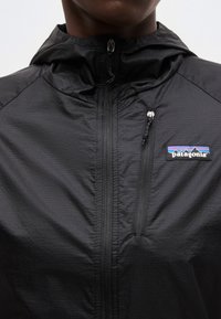 Sort Patagonia hættetrøje med frontlynlås og lomme med lynlås på brystet, båret af en person vist fra hagen til brystet.