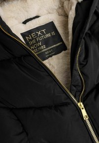 Schwarze Pufferjacke mit einem goldenen Reißverschluss und einem Etikett mit der Aufschrift "NEXT". Das Innenfutter ist mit plüschigem, hellfarbigem Kunstfell ausgekleidet. Sauberes Design, ohne Muster.