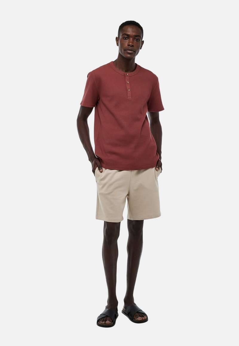 Chemise rouge à rayures côtelées à manches courtes avec fermeture à boutons, associée à un short beige. Des sandales noires complètent la tenue. Fond uni.